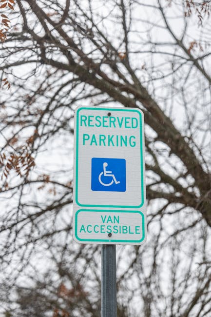 A close-up view of a white reserved parking sign featuring a blue wheelchair symbol, indicating a designated disabled parking space. The sign has green borders and text, with the words 'RESERVED PARKING' at the top and a green rectangular section at the bottom reading 'VAN ACCESSIBLE.' The sign is mounted on a grey metal pole positioned outdoors against a background of leafless tree branches and an overcast sky, suggesting a winter or early spring season. This type of signage is relevant for home relocation services, such as those provided by Man with Van Harlesden, to facilitate efficient furniture transport and loading processes during house removals in compliance with local regulations, like Brent Council's rules for skip permits and house clearances.