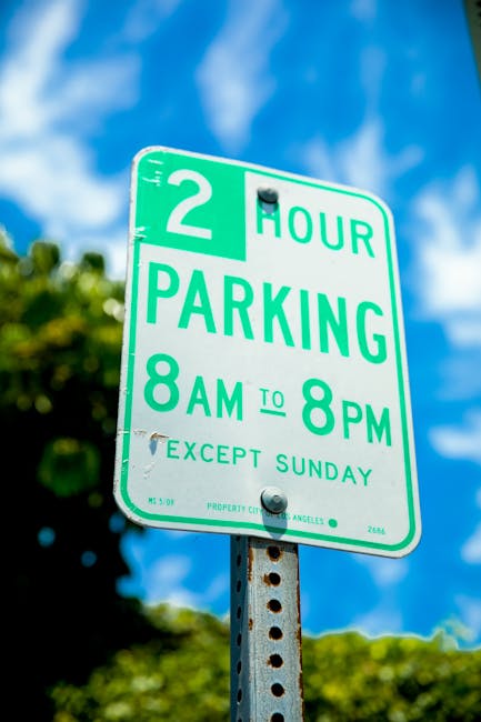 Close-up of a green and white parking restriction sign mounted on a metal pole, indicating a 2-hour parking limit from 8 am to 8 pm, except on Sundays. The sign features bold green text on a white background with the words '2 HOUR PARKING' and details about timing and exceptions. The background shows a partly cloudy blue sky with some blurred green foliage, suggesting an outdoor urban or residential area. The sign is relevant to house removals and moving logistics, indicating parking regulations that may impact the loading and unloading process during home relocations, which companies like Man with Van Harlesden need to consider.