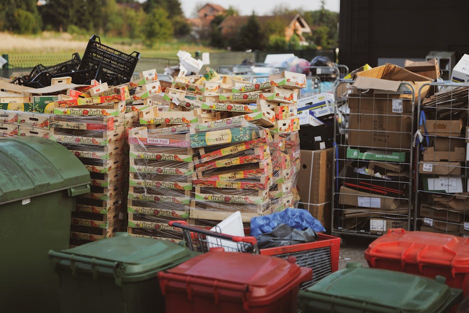 Outdoor area showing multiple stacks of flattened cardboard boxes with fruit branding, arranged in front of a black plastic crate and a metal shelving unit filled with various boxes and packaging materials. Several large green and red plastic waste bins are positioned in the foreground, while a red shopping trolley and some scattered packing supplies are visible on the ground. The environment suggests a preparation or collection site for furniture transport and packing during a home relocation or removal process, with natural lighting indicating daytime. The scene illustrates the handling and organization of packing materials, which are integral steps in furniture transport and moving logistics services offered by Man with Van Harlesden.
