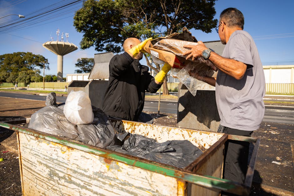 Two men are engaged in a home relocation task involving the disposal of bulky waste materials in an outdoor area. One man, wearing a black jacket and yellow gloves, is lifting and placing a cardboard box filled with miscellaneous items into a large, rusty metal skip, which contains several black and grey plastic rubbish bags and other waste. The other man, dressed in a grey t-shirt and black trousers, is assisting with the loading process, holding the box steady. The setting is outdoors on a paved surface near a street, with a background featuring trees, a tall water tower, utility poles, and a clear blue sky. Natural sunlight illuminates the scene, highlighting the effort involved in loading waste during a house removal or furniture transport process. This image reflects professional waste disposal and clearance services that may be part of a comprehensive move managed by Man with Van Harlesden, with a focus on efficient and safe disposal of unwanted bulky items during the packing and moving stages.