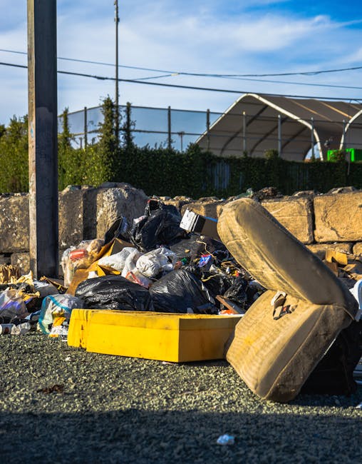 Outdoor area showing multiple stacks of flattened cardboard boxes with fruit branding, arranged in front of a black plastic crate and a metal shelving unit filled with various boxes and packaging materials. Several large green and red plastic waste bins are positioned in the foreground, while a red shopping trolley and some scattered packing supplies are visible on the ground. The environment suggests a preparation or collection site for furniture transport and packing during a home relocation or removal process, with natural lighting indicating daytime. The scene illustrates the handling and organization of packing materials, which are integral steps in furniture transport and moving logistics services offered by Man with Van Harlesden.