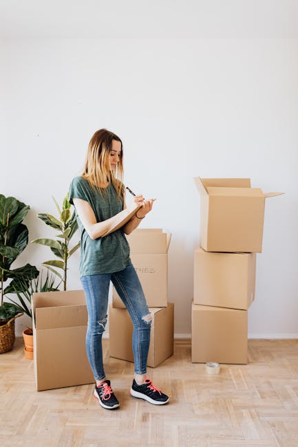 A young woman with shoulder-length blonde hair dressed in a casual green T-shirt, ripped blue jeans, and black athletic shoes, standing indoors on a wooden floor during a home relocation process. She is holding a notepad and pen, inspecting and taking notes on moving boxes, which include an open cardboard box on the floor and a larger, sealed box behind her. To her left, a tall green potted plant with broad leaves sits against a plain white wall. Several packed and partially assembled cardboard boxes are stacked to her right, with one slightly open showing cardboard flaps. The scene captures a typical packing and moving activity, with the woman reviewing or planning her move, supported by visible packing materials and the interior environment suitable for house removals by companies like Man with Van Harlesden.