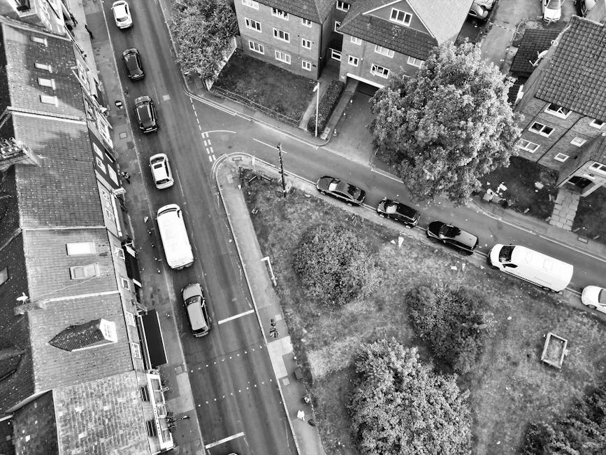 An aerial black-and-white photograph showing a residential street with parked cars along both sides, adjacent to a small green space with trees and bushes. The street has houses with pitched roofs and brick facades lining the road, with some entering driveways. A white van from Man with Van Harlesden is parked at the curb on the right side, near the entrance to a property. In the foreground, a loading process is visible with cardboard boxes and wrapped furniture being carried, possibly into or out of the van. Moving blankets and straps are seen securing items during the load or unload. The environment is well-lit, with shadows cast by trees and vehicles, highlighting the urban setting typical for house removals, packing, and furniture transport services involved in home relocation or same-day moving logistics.