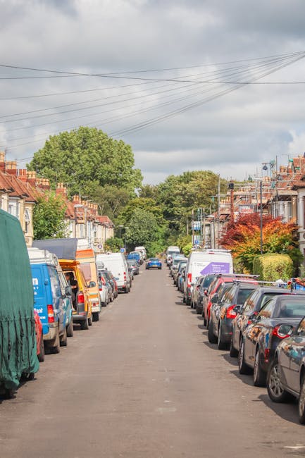 A residential street scene with parked cars lining both sides of the narrow tarmac road, which features a central lane. Vehicles include a range of small to medium-sized cars, vans, and delivery trucks, some with protective coverings or open rear doors. In the background, a house with a pitched roof and brick chimney is visible on the left, while a large green tree provides partial shade at the end of the street. Overhead, multiple power lines cross the cloudy sky. The environment appears calm and typical of suburban housing areas, suitable for home relocation and furniture transport activities. Occasionally, vans with company branding linked to Man with Van Harlesden can be seen parked along the pavement, indicating potential relocation services in progress or ready for assistance from the removals company. The scene captures the typical urban setting where loading and unloading processes for home moving services could take place, with no visible people or loading activity in this image.