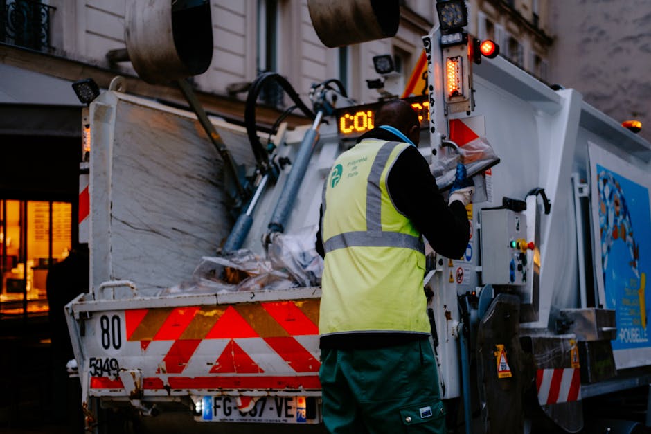 A close-up of a person wearing orange protective overalls and white gloves, holding a blue plastic bag filled with waste material. The individual appears to be involved in waste disposal or removal ac