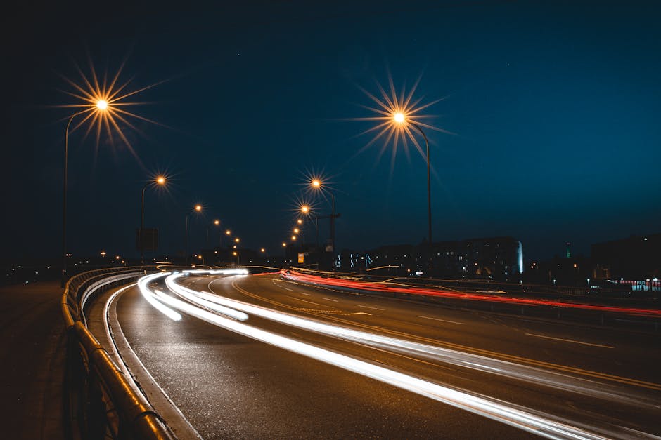 Nighttime scene of a curved urban road illuminated by evenly spaced streetlights, casting a warm yellow glow against a dark blue sky. Light trails from moving vehicles with white headlights and red taillights create streaks across the road, indicating long exposure photography. The road features a metal guardrail on the left side, and in the background, faint outlines of buildings are visible with some illuminated windows. The scene suggests a busy transport route, relevant to the context of house removals and transportation logistics, as conducted by companies like Man with Van Harlesden, especially during peak relocation times near Willesden Junction.