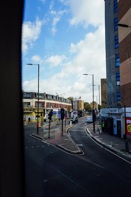 View of a city street scene captured from inside a building, featuring a curved road with tram tracks, streetlights, and a bus stop with a yellow shelter on the left side. Pedestrians are walking along the pavement, and several vehicles, including a white van, are parked or moving nearby. The background shows a mix of modern and older multi-storey buildings, some with shopfronts or advertisements, under a partly cloudy sky with patches of blue. The image conveys an urban environment suitable for house removals or furniture transportation, as seen from the perspective of a moving company preparing equipment or planning logistics near Willesden Junction, with [COMPANY_NAME] providing removals services in the area.