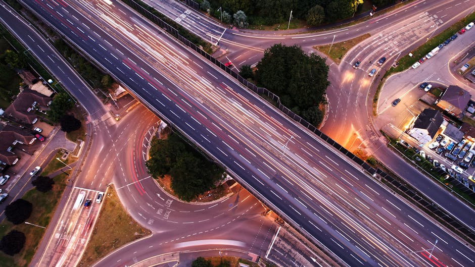 Nighttime scene of a curved urban road illuminated by evenly spaced streetlights, casting a warm yellow glow against a dark blue sky. Light trails from moving vehicles with white headlights and red taillights create streaks across the road, indicating long exposure photography. The road features a metal guardrail on the left side, and in the background, faint outlines of buildings are visible with some illuminated windows. The scene suggests a busy transport route, relevant to the context of house removals and transportation logistics, as conducted by companies like Man with Van Harlesden, especially during peak relocation times near Willesden Junction.