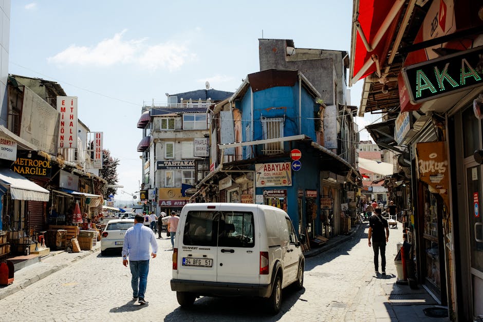 A narrow street in Harlesden High Street showing a typical urban environment with small retail shops, signage, and pedestrians. In the foreground, a white van with its rear doors closed is parked on the cobbled pavement, with a man walking nearby. To the right, the exterior of a shop displays various signs and awnings, including a green LED sign. Adjacent to the van, a man in a light blue shirt and jeans is walking along the street, while another individual dressed in black is further down. The buildings lining the street are multi-story, with visible balconies and signage indicating different businesses such as a pharmacy, cafes, and other retail outlets. Several cardboard boxes, plastic packaging, and moving supplies are visible near the shops, suggesting ongoing packing or loading activities typical of home relocation or furniture transport. The scene captures the vibrant, busy atmosphere of urban moving logistics in Harlesden, with natural daylight illuminating the area and a clear sky overhead.