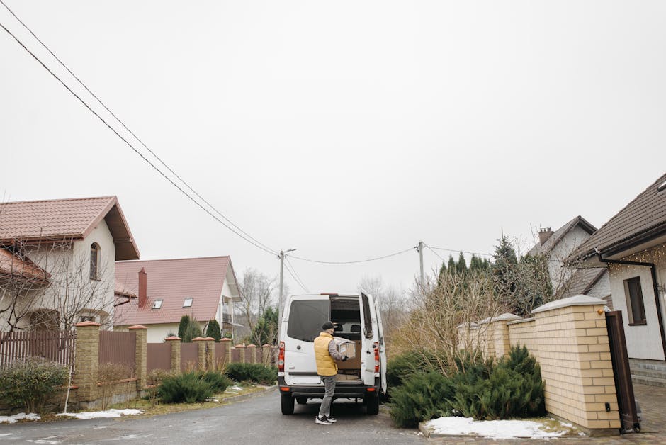 A man wearing a yellow jacket and black beanie is unloading a cardboard box from the open rear doors of a white van parked on a residential street in Harlesden. The image shows the van positioned on t