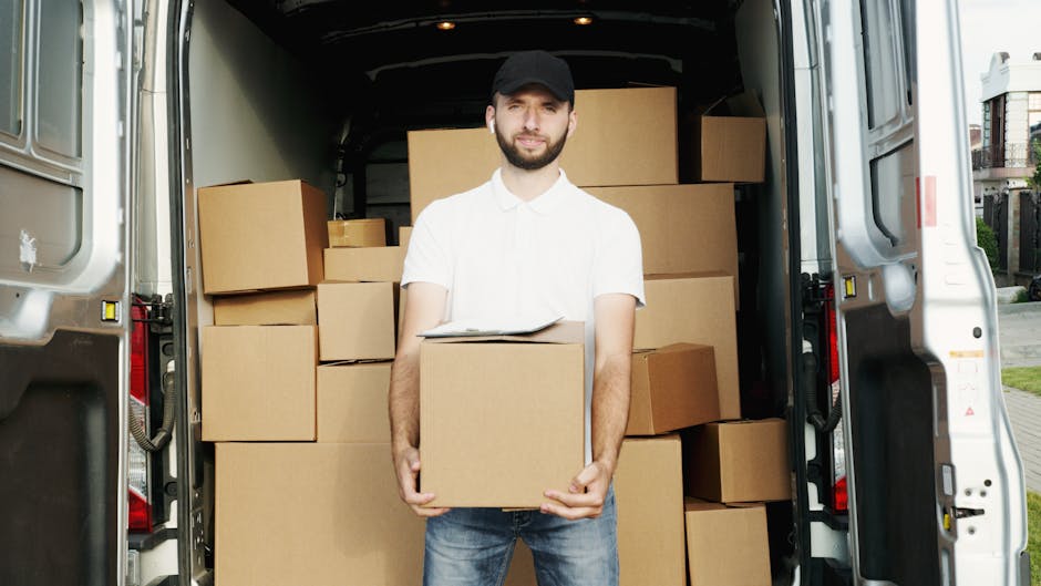 A man wearing a yellow jacket and black beanie is unloading a cardboard box from the open rear doors of a white van parked on a residential street in Harlesden. The image shows the van positioned on t