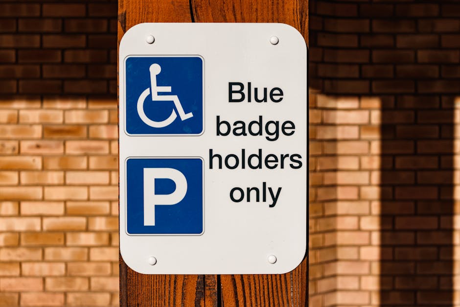 A white rectangular parking sign mounted on a wooden post in front of a brick wall. The top left corner of the sign displays a blue symbol of a person in a wheelchair, indicating accessibility for disabled badge holders. To the right of this symbol, the sign reads 'Blue badge holders only' in black text. Beneath the text, there is a large blue square with a white letter 'P', denoting parking. The sign's positioning suggests it is situated outside a property, possibly near an entrance or parking area used for home relocation or furniture transport. The background features a neatly arranged brick wall, and the lighting indicates daylight, with a shadow cast on the brick surface. This type of signage relates to parking restrictions and permits that can impact moving logistics managed by companies like Man with Van Harlesden, especially when planning a house removal or furniture transport in Harlesden.
