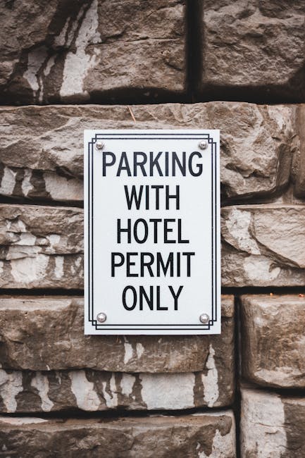 A rectangular white metal sign with black text reading 'PARKING WITH HOTEL PERMIT ONLY' is mounted on a rough brown stone wall using four screws, one at each corner. The wall features large, uneven stones with visible mortar lines, creating a textured surface. The sign is positioned at eye level, indicating restrictions for parking in the area. The setting appears to be outside a building in Harlesden, relevant to local removals and house relocation services offered by Man with Van Harlesden. In the background, part of a moving process may involve loading furniture, boxes, and household items into a van for home relocation, with the sign implying parking regulations that could impact the logistics of the move.
