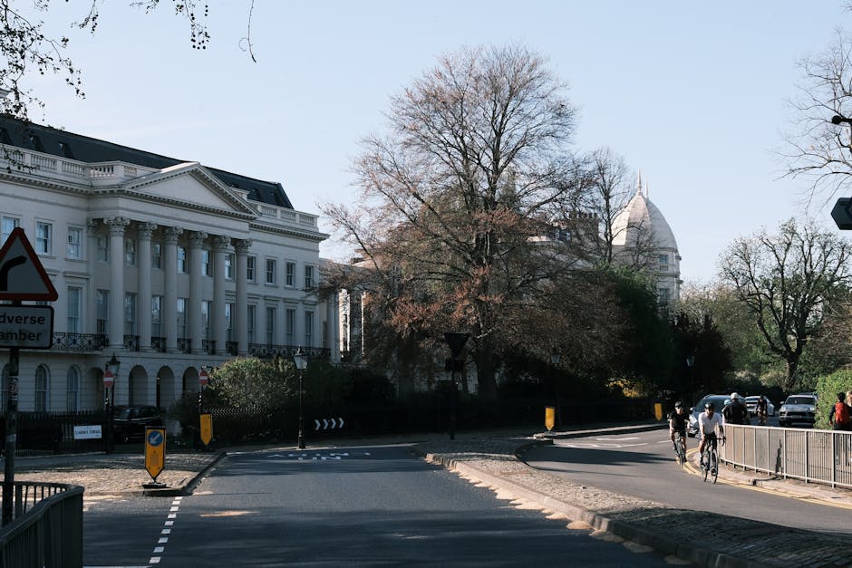 A street scene showing the entrance to a property with a paved driveway and a white, neoclassical-style building with tall columns and decorative elements in the background, situated in a mature residential area during daylight. To the right, there are pedestrians walking and cyclists riding along the pavement, with a metal railing separating the pedestrians from the street. Several parked cars are visible near the building, which is partially shaded by large, leafless trees indicating an early spring or late autumn setting. The scene includes a traffic sign indicating an adverse chamber, and the road features small road blocks or bollards guiding vehicle and pedestrian movement. This image captures a typical urban environment suitable for home relocation or furniture transport activities, with an emphasis on the well-maintained street, parking area, and surrounding greenery, relevant to the services offered by Man with Van Harlesden.