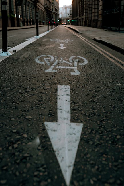 A close-up view of a designated cycling lane in an urban environment, with white painted symbols and arrows on the asphalt indicating the direction for cyclists. The lane is separated from the main road by a row of black bollards with reflective strips, and features a bike lane symbol near the bottom of the image. In the background, there are tall buildings on either side of the street, with some traffic lights visible at the intersection ahead. The scene is captured under natural daylight, highlighting the texture of the asphalt and the clarity of the road markings, which are part of the street infrastructure supporting safe cycling and transportation, relevant to logistics and moving services such as those offered by Man with Van Harlesden, especially when planning routes for home relocations or furniture transport.