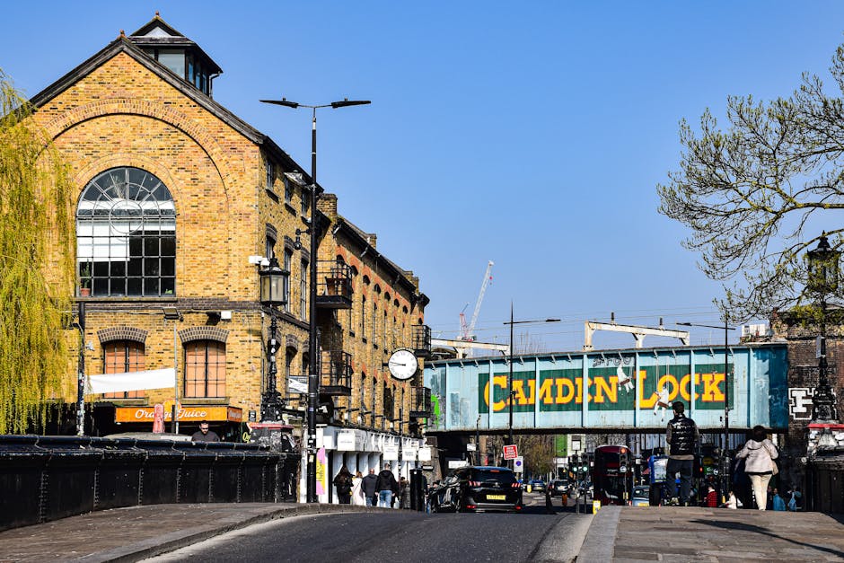 A street scene in Harlesden featuring a large, historic brick building with a prominent arched window on the upper floor, and a modern railway bridge with yellow and green signage reading 'Camden Lock