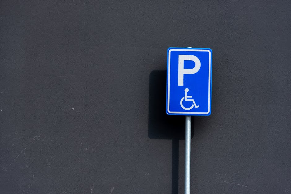 A blue parking sign with a white 'P' and a wheelchair symbol indicating disabled parking, mounted on a silver metal pole against a dark grey textured wall. The sign casts a slight shadow to the left, with even lighting suggesting daylight conditions. The background wall is plain without additional markings, providing clear visibility of the sign. This parking sign is situated outdoors, likely nearby a building entrance or designated parking area. It is positioned at a height accessible to drivers and visitors, supporting accessible parking for individuals with disabilities, which is relevant for considerations around residential or commercial relocations managed by Man with Van Harlesden. The clean, simple design of the sign contributes to easy recognition for drivers seeking accessible parking spaces during house removals or furniture transport processes.