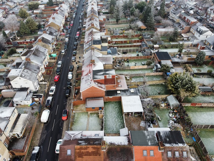 An aerial view of a suburban residential street during daytime, showing rows of terraced and semi-detached houses with pitched roofs, some covered with snow. Cars are parked along both sides of the asphalt road, which runs through the centre of the scene. Behind the houses, there are private backyards with various fences, sheds, patios, and gardening equipment. The backyards contain a mix of lawn areas, paved sections, and storage sheds made of wood or metal, some with plastic covers. The environment appears to be in winter, with leafless trees and snow on rooftops and yards. The scene captures a typical urban environment where house removals and furniture transport might take place, with some yards showing signs of recent activity, such as packed boxes or furniture, indicating ongoing packing or moving preparations. The image provides context relevant to home relocation services like those offered by Man with Van Harlesden, illustrating the practical setting for a loading process or moving logistics in a residential area.